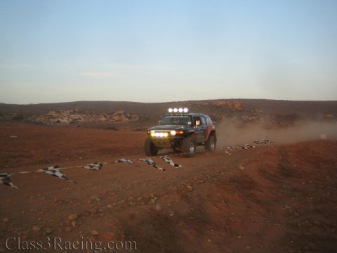 1990 Ford Bronco - off-road race vehicle with a roof light bar and headlights on crosses rocky reddish desert terrain at dusk.