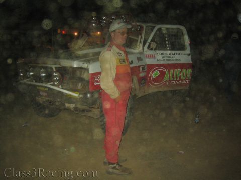 1990 Ford Bronco - man in a red racing suit stands beside a white California Tomatoes off-road race truck in a dusty night pit.