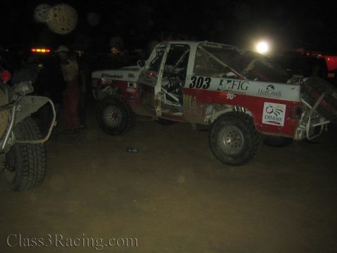 1990 Ford Bronco - red and white off-road race truck number 303 with FIG and DiMare sponsor decals sits in a dim dusty night pit.