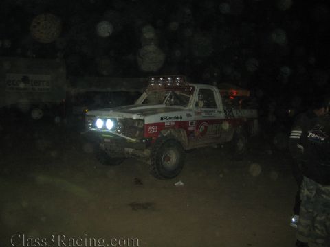 1990 Ford Bronco - white and red off-road race truck number 303 shines its headlights in a dark night pit as a person stands at right.