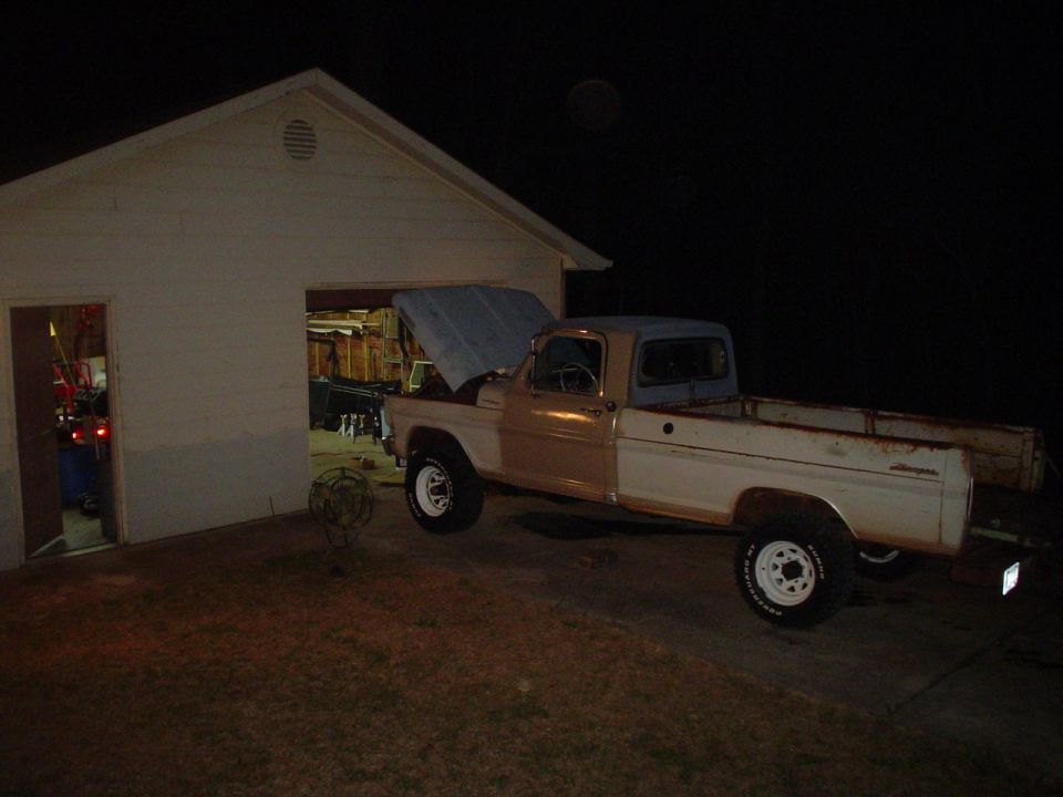1967 Ford F-250 pickup truck with hood open parked next to a garage at night