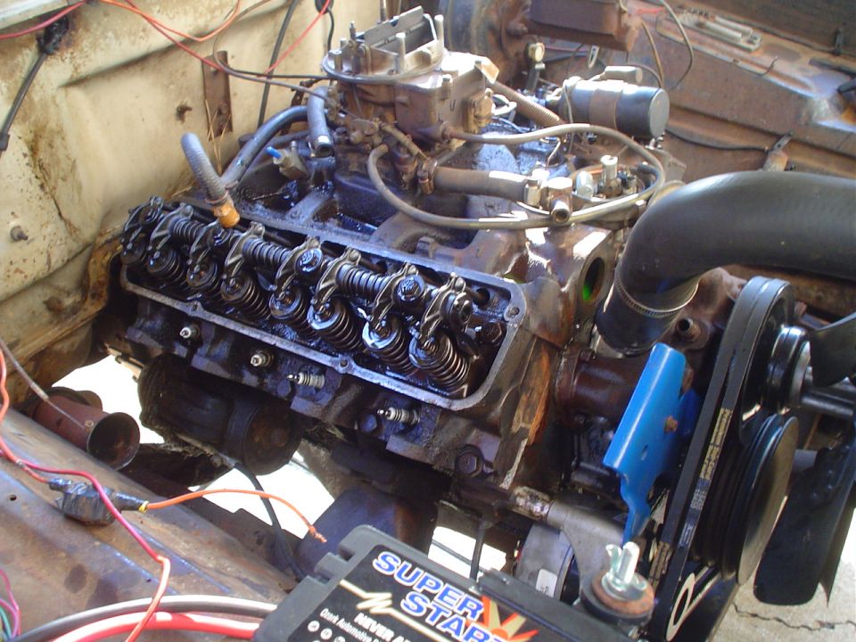 Close-up of a V8 engine in a vehicle, showing valve covers, carburetor, and belts. A Super Start battery is in the foreground
