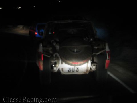 1990 Ford Bronco - blurry low-light rear view of an off-road truck carrying a spare tire with number 303 driving on a dark road.