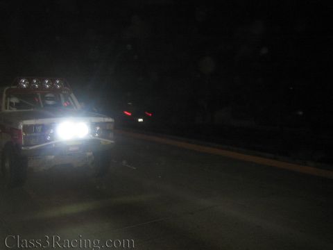 1990 Ford Bronco - white off-road race truck with roof lights and glaring headlights sits at the edge of a dark road at night.