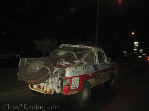 1990 Ford Bronco - Rear three-quarter view of red and white race truck number 303 with mounted spare tires driving a road at night.
