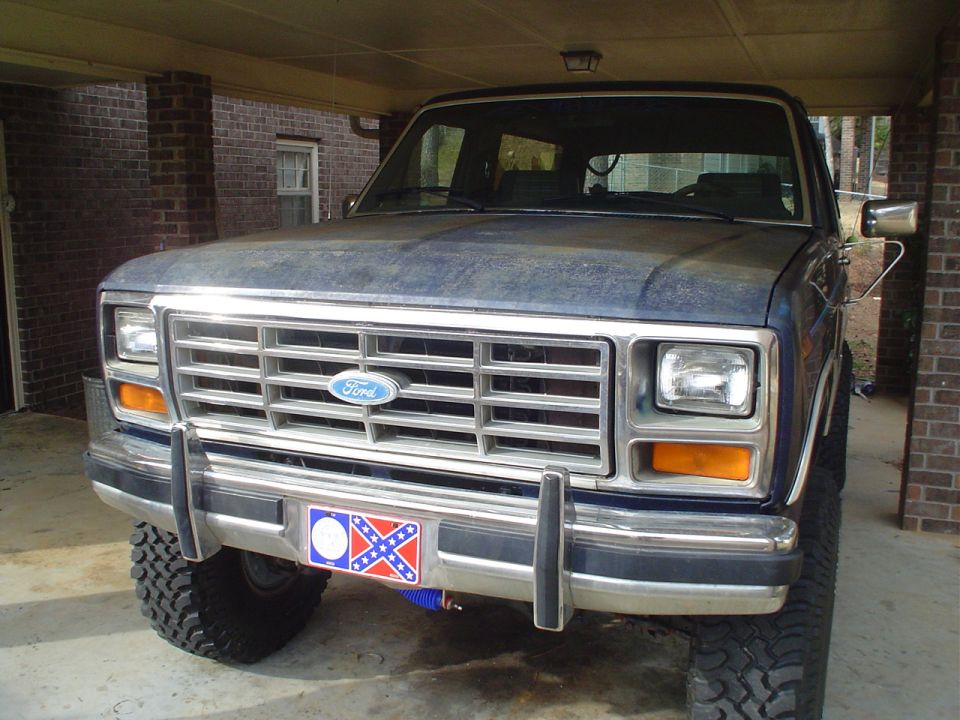 Blue Ford Bronco front view, showing grille and headlights. License plate frame has a Confederate flag