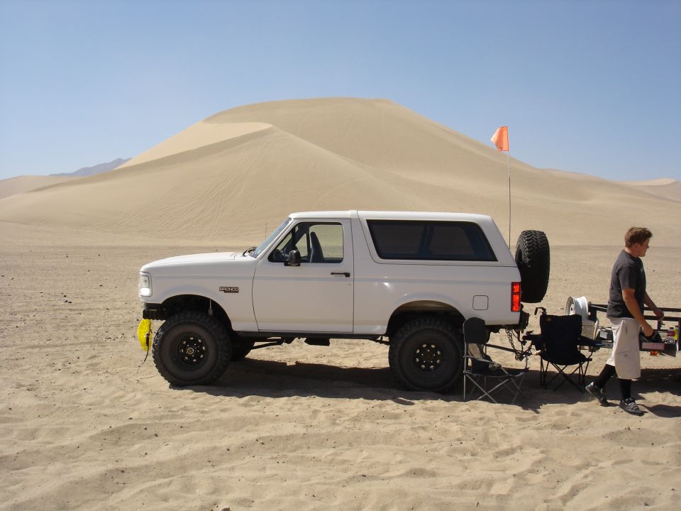 White 1996 Ford Bronco with aftermarket wheels parked in sand dunes, side view