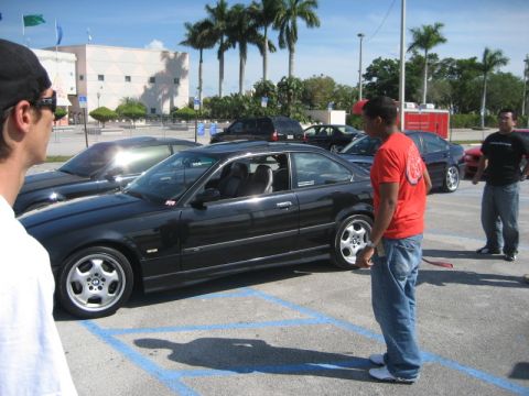 1984 BMW 3 Series - A black BMW coupe is parked in a lot, with several people standing nearby. Another car is visible to the left.