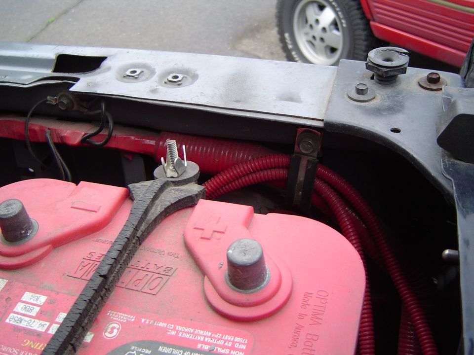 Engine bay with red Optima battery and red wiring conduit. Another vehicle is partially visible