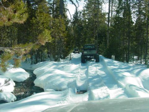 1986 Ford Bronco - Off-road vehicles navigate a snowy trail through a wooded area with a stream alongside.
