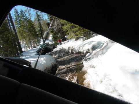 1986 Ford Bronco - Two vehicles are stuck in a snowy, muddy ditch in a wooded area, with one vehicle partially submerged in water.