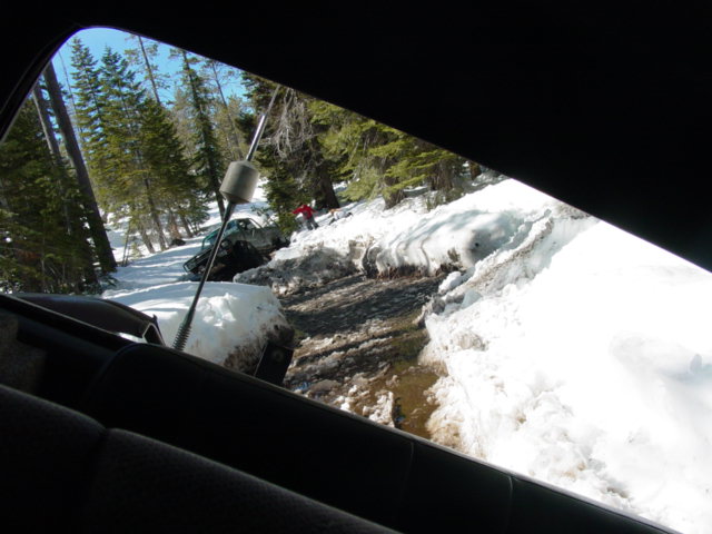 Two off-road vehicles are stuck in a snowy, muddy ditch in a forest. One vehicle is partially submerged in water