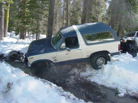 1986 Ford Bronco - A two-tone Ford Bronco is stuck in a muddy, snowy rut in a wooded area, with another vehicle visible behind it.
