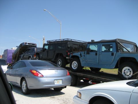 1986 Ford Bronco - A car carrier transports multiple vehicles, including two Humvees and a silver coupe, under a clear blue sky.