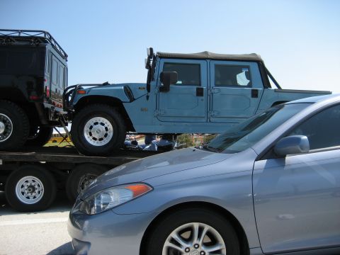 1986 Ford Bronco - A blue Hummer H1 is transported on a trailer, partially obscured by a silver sedan.