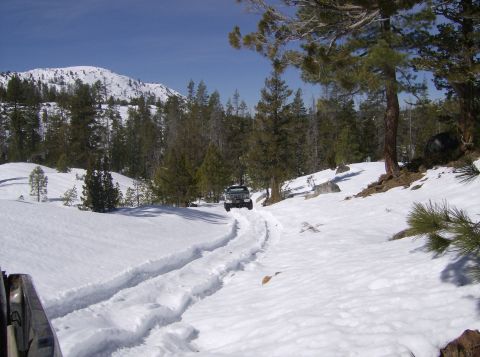 1986 Ford Bronco - A vehicle navigates a snow-covered trail with tire tracks leading up a hill towards a wooded area.
