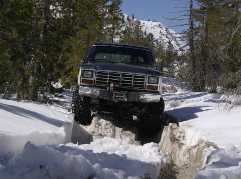 1986 Ford Bronco - A lifted Ford Bronco navigates a snowy mountain trail, its front grille and winch clearly visible.