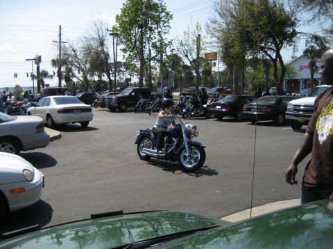 1986 Ford Bronco - A rider cruises through a parking lot filled with cars and motorcycles on a sunny day.