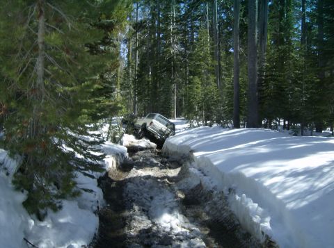 1986 Ford Bronco - A white SUV is stuck in a snowy ditch on a wooded road, with trees and snowbanks surrounding it.