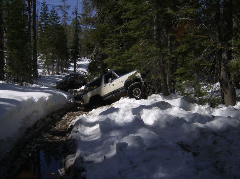 1986 Ford Bronco - Two off-road vehicles navigate a snowy, wooded path, with one appearing stuck.
