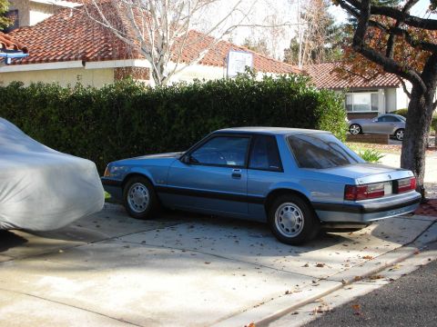 1990 Ford Mustang - A blue 1990 Ford Mustang coupe is parked on a driveway next to a car covered with a tarp.