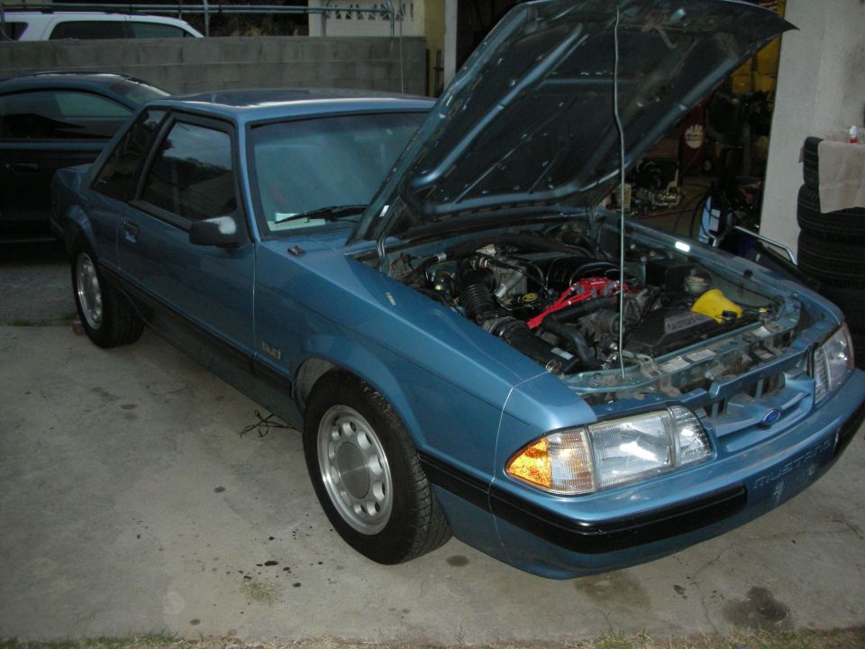 1990 Ford Mustang coupe, blue, hood open revealing engine bay. Another car is partially visible in the background