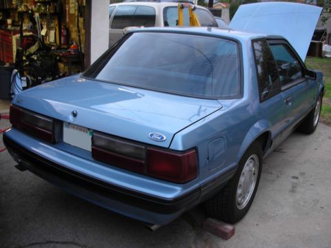 1990 Ford Mustang - A blue 1990 Ford Mustang is parked in front of a garage, with its hood open.