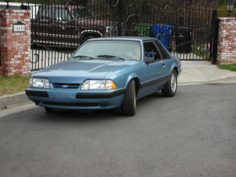 1990 Ford Mustang - This blue 1990 Ford Mustang coupe is seen parked in front of a gated driveway, with another vehicle visible behind the gate.