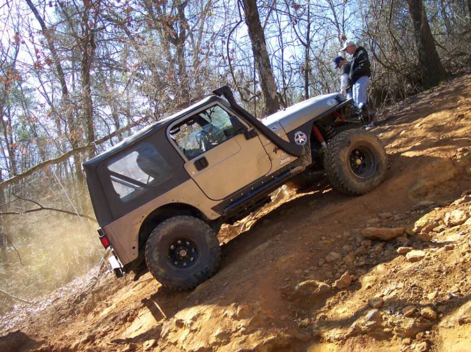 Tan Jeep Wrangler with black soft top and snorkel driving up a dirt hill, two people standing nearby