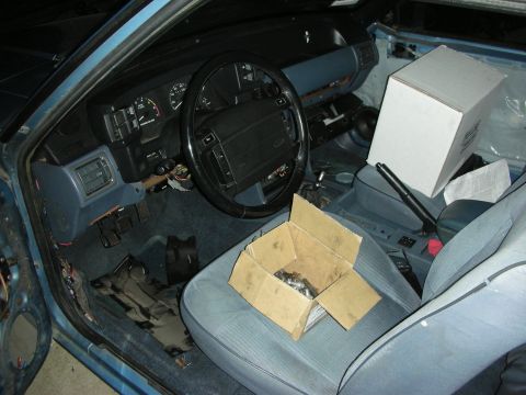 1990 Ford Mustang - Bare interior of a vehicle under restoration with blue-gray cloth seats, cardboard parts boxes on seat, dash intact.