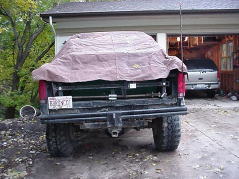 1992 Ford Bronco - Rear view of a lifted full-size Ford Bronco parked on a gravel driveway with a tan tarp draped over the cab and cargo area. A vanity plate…