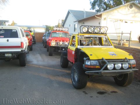 1990 Ford Bronco - yellow off-road race truck with a roof light bar sits in a driveway beside red and white trucks at a suburban home.