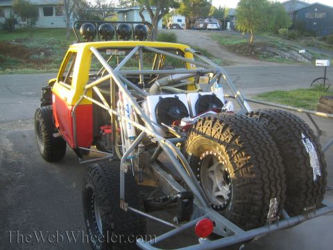 1990 Ford Bronco - Rear view of a yellow-and-red tube-chassis off-road race truck with mounted spare tires and exposed roll cage parked in a driveway.