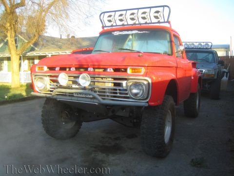1990 Ford Bronco - lifted vintage orange Ford pickup with roof light bar and oversized tires parked beside another truck on pavement.