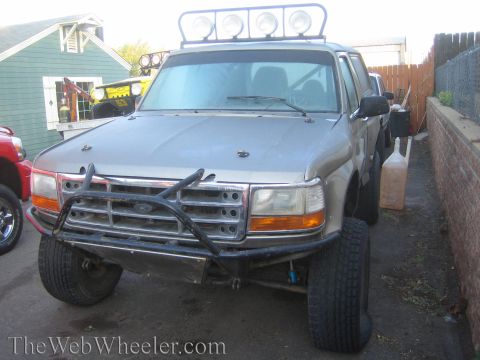 1990 Ford Bronco - silver 1990s Ford pickup fitted with a roof light bar and tubular front bumper parked in a narrow side yard.