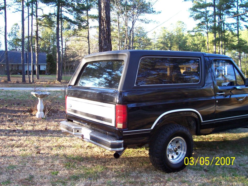 Black two-door Ford Bronco SUV, rear 3/4 view, parked on grass with chrome bumper and mud tires