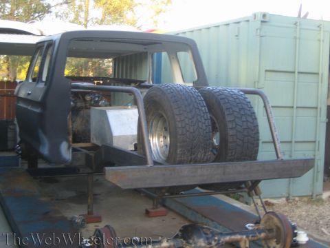 1990 Ford Bronco - bare dark truck cab on stands with a fabricated rear frame, dual rear tires, and exposed solid axle in a yard.