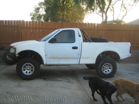 1990 Ford Bronco - Side view of a white lifted Ford pickup project with no front fender in a backyard, with a black and a tan dog nearby.