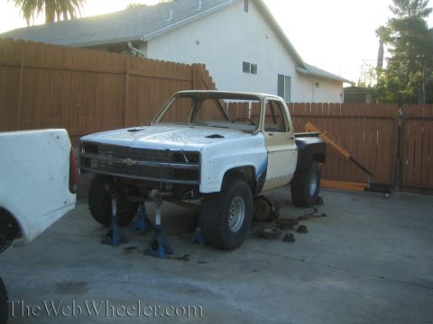 1990 Ford Bronco - white Chevrolet stepside pickup project up on jack stands with removed parts on the ground in a residential driveway.
