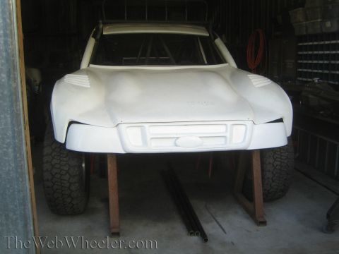 1990 Ford Bronco - Front view of a smooth white fiberglass off-road truck body shell with a Ford emblem, mounted on stands in a metal shed.