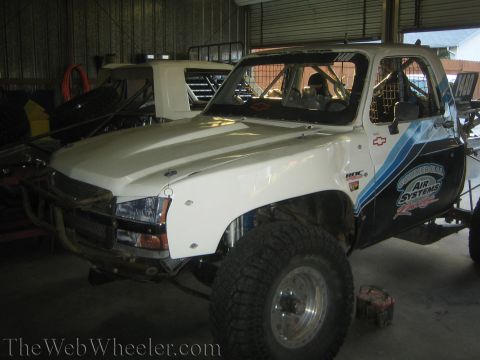1990 Ford Bronco - white Chevrolet prerunner truck with roof spoiler, tube front bumper, and Air Systems livery parked inside a metal shed.