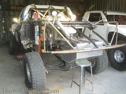 1990 Ford Bronco - Front-end tube-chassis race truck build showing coilover shocks, fuel cell, and a stool inside a corrugated-metal shed.