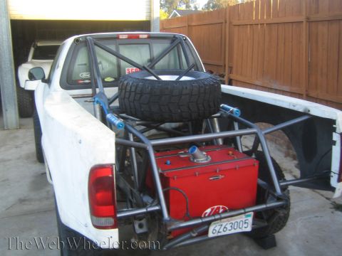 1990 Ford Bronco - Rear bed of a white Ford prerunner truck holding a spare tire, red fuel cell, and tubular bracing in a driveway.