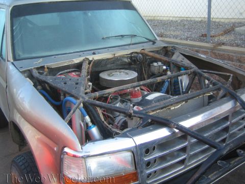 1990 Ford Bronco - Engine bay of a project truck showing a V8 with chrome air cleaner, blue wiring, radiator, and exposed tubular grille framing.