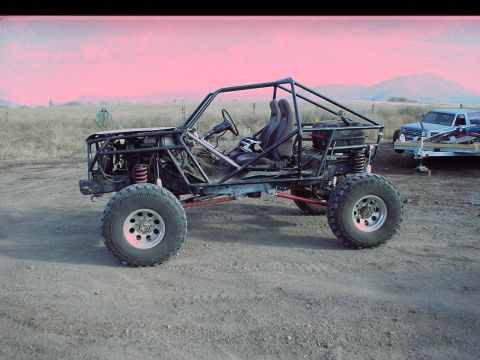 1984 Ford Bronco - Custom buggy with roll cage and racing seats sits on dirt. Another vehicle is on a trailer behind it.
