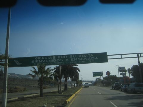 1990 Ford Bronco - green overhead sign reading "Gracias por su visita / Bienvenidos a Ensenada" spans a sunlit road lined with palm trees.