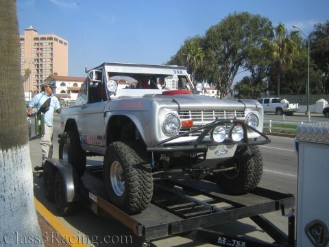 1990 Ford Bronco - silver lifted vintage Ford Bronco off-road race truck sits on a flatbed trailer near a beachfront hotel and palm trees.