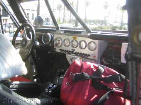 1990 Ford Bronco - stripped interior of an off-road race truck reveals a roll cage, a panel of round gauges and switches, and a red racing seat.