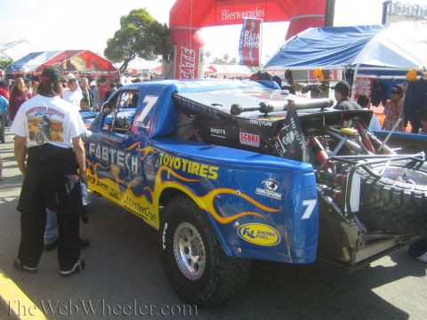 1990 Ford Bronco - blue number 7 Fabtech / Toyo Tires off-road race truck sits in a crowded pit area beneath a Baja event arch and tents.