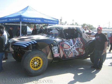 1990 Ford Bronco - black and silver number 54 off-road buggy with skull and "Choppers" graphics parked by a Fox Racing Shox tent, a man posing beside it.